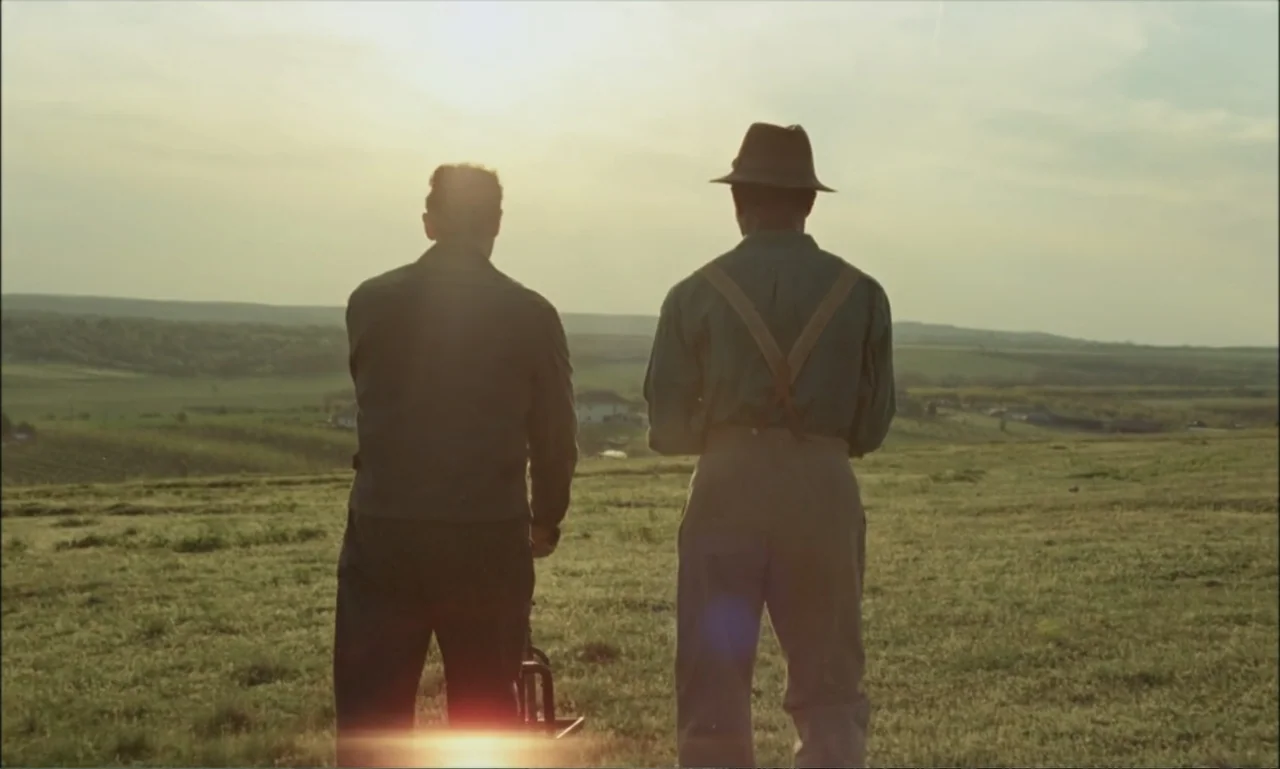 The Brutalist (2024) movie still: fullbody — An open grassy field stretches toward a distant tree line under a bright sky. Two men stand with their…