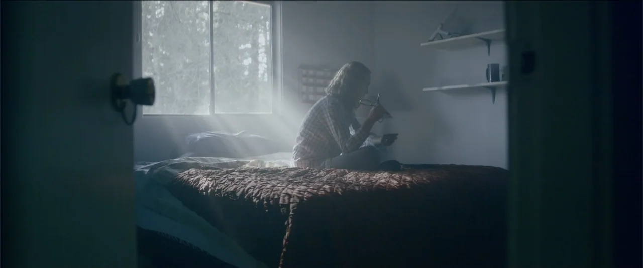 The Hollow Child (2018) movie still: medium — A dimly lit room contains a bed with a quilted brown cover and window light. A woman sits on the edge…