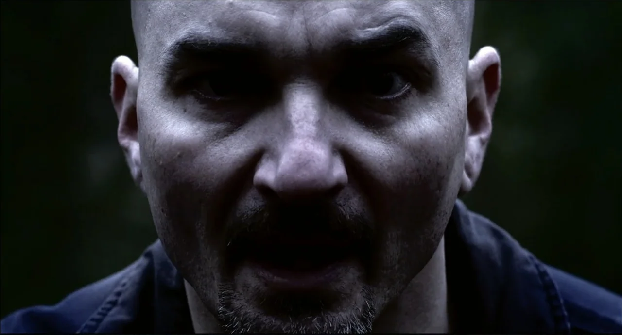The Secret Village (2013) movie still: closeup — A dark space serves as the backdrop for a close portrait of a man. The man looks directly into the…