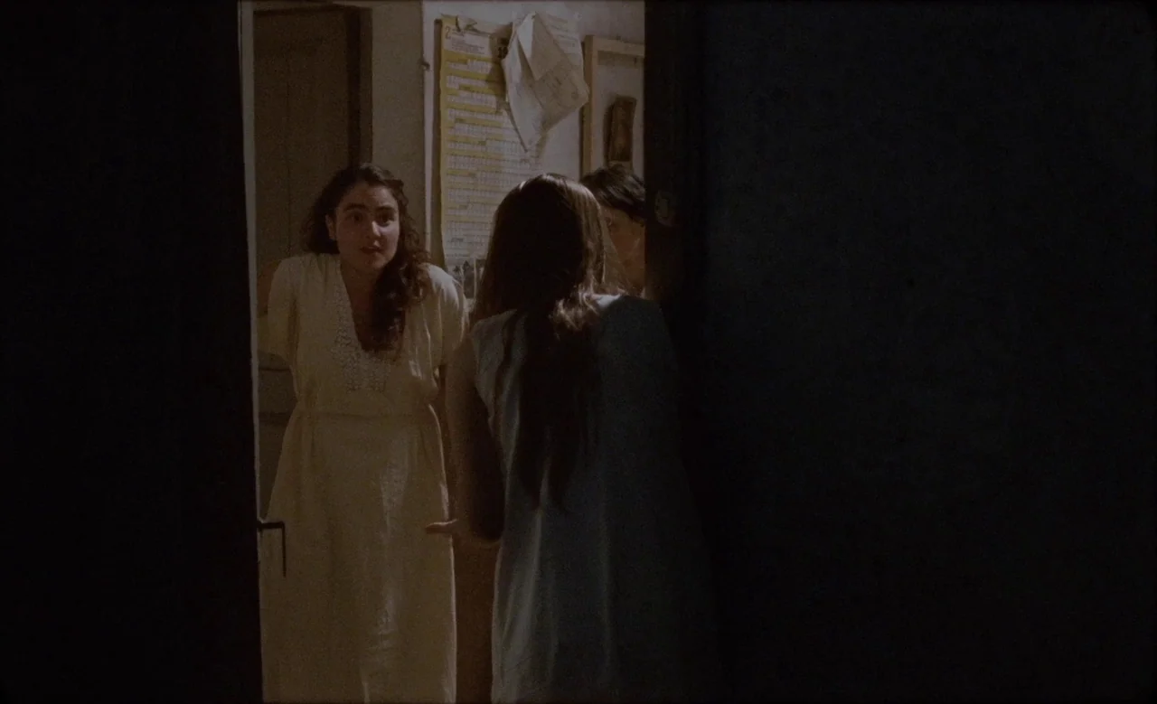 Happy as Lazzaro (2018) movie still: medium — A dimly lit home interior features two women conversing across a narrow doorway. A wall calendar hangs…