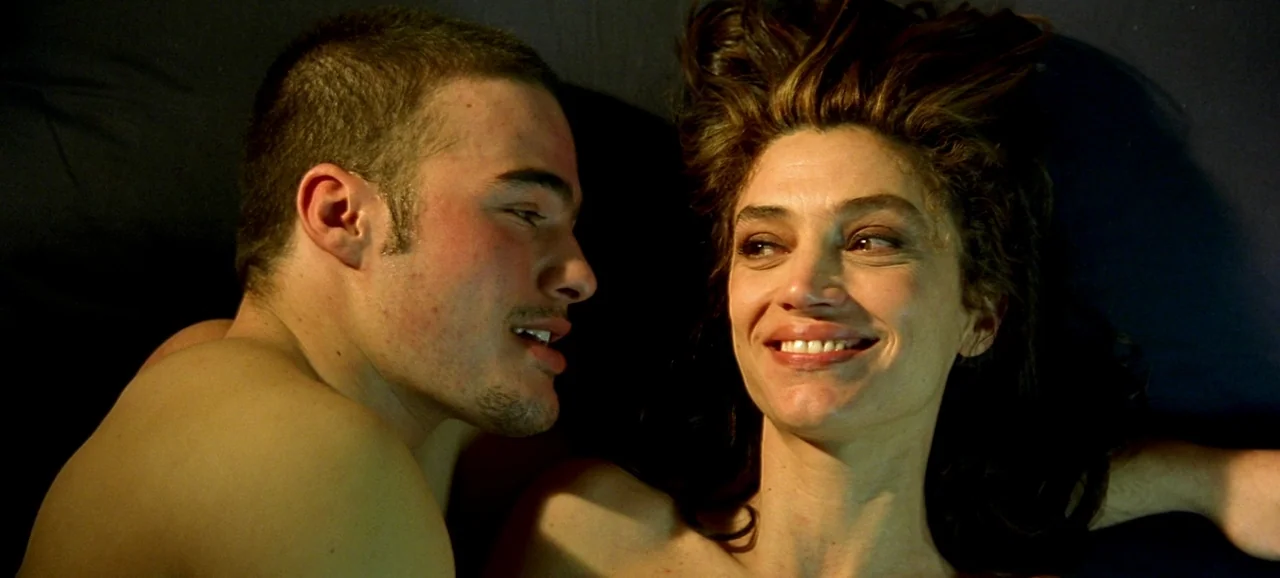 Live Flesh (1997) movie still: closeup — A dark space with two individuals lying close together on a flat surface. A man with short hair gazes at a…