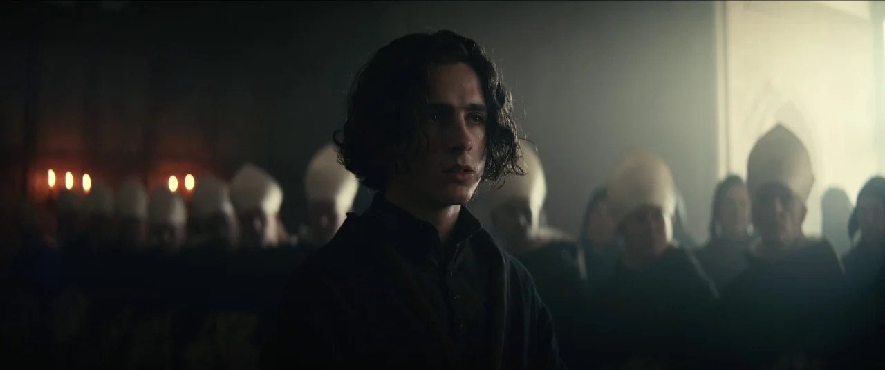 The King movie still: medium — A stone interior space features a crowd of figures wearing white pointed head coverings. A young man stands in the…