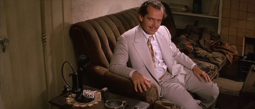 The Fortune (1975) movie still: medium — A 1930s-style room features a brown velvet sofa and a vintage telephone table. A man in a light suit sits…