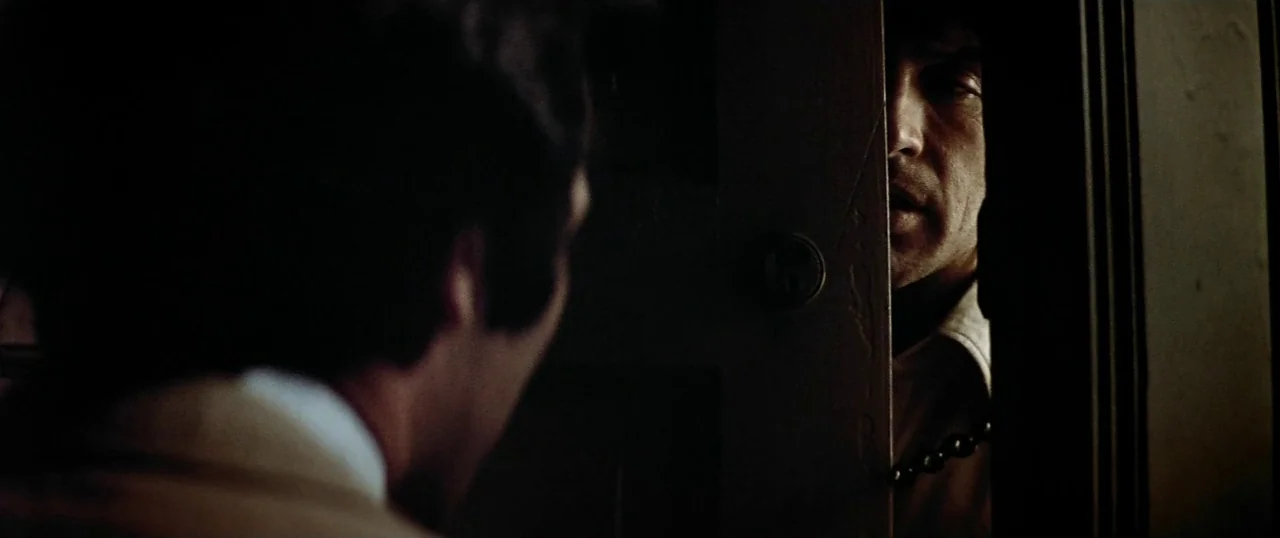 The Parallax View (1974) movie still: medium — A man stands behind a partially opened door in a dimly lit space. The other man is visible in the…