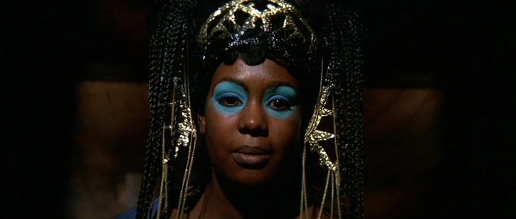 Fellini – Satyricon (1969) movie still: closeup — A woman stands in a darkened space wearing ornate jewelry and bold makeup. She looks directly…