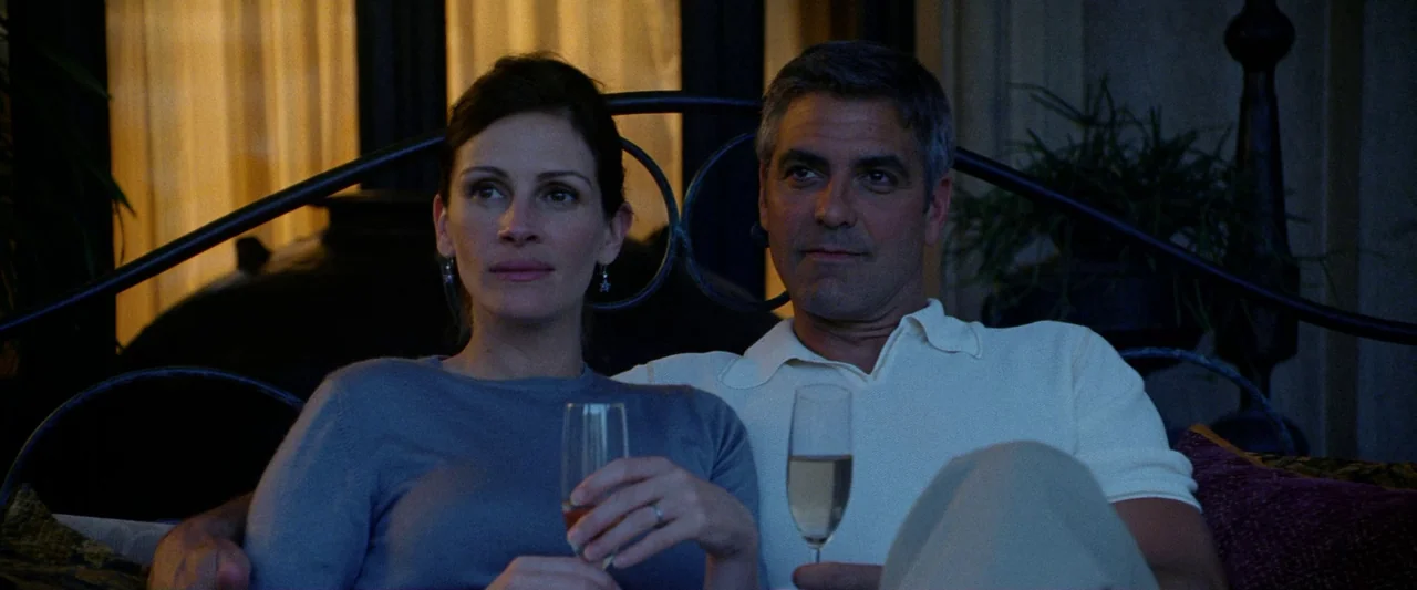 Ocean’s Twelve (2004) movie still: medium — A man and woman sit side by side on an outdoor patio furniture set. They hold glass flutes filled with…