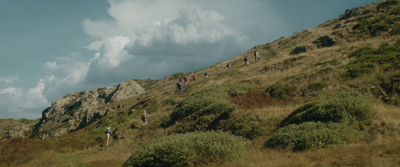 Beast (2017) movie still: wide — A grassy hillside leads upward to a ridgeline under a sky with large clouds. A group of hikers travels along a dirt…