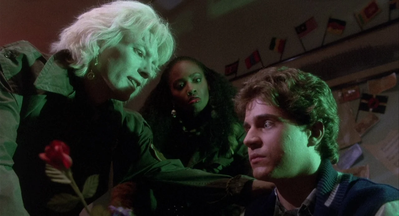 Vamp (1986) movie still: medium — A dimly lit room features walls decorated with small international flags hung on strings. Two women lean toward a…