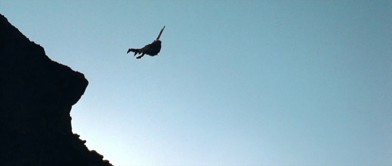 The Hills Have Eyes (2006) movie still: wide — A dark rocky ledge projects into a wide expanse of pale blue sky. A solitary figure freefalls through…