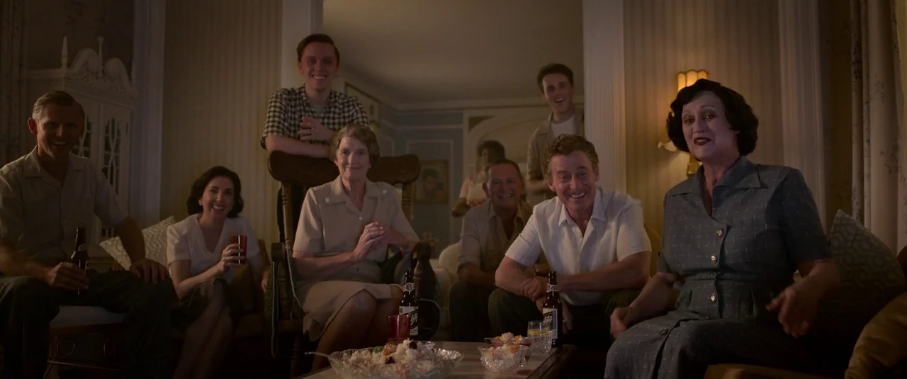 Elvis (2022) movie still: wide — A dimly lit 1950s living room features a group of adults gathered together. The group poses for a photograph behind…