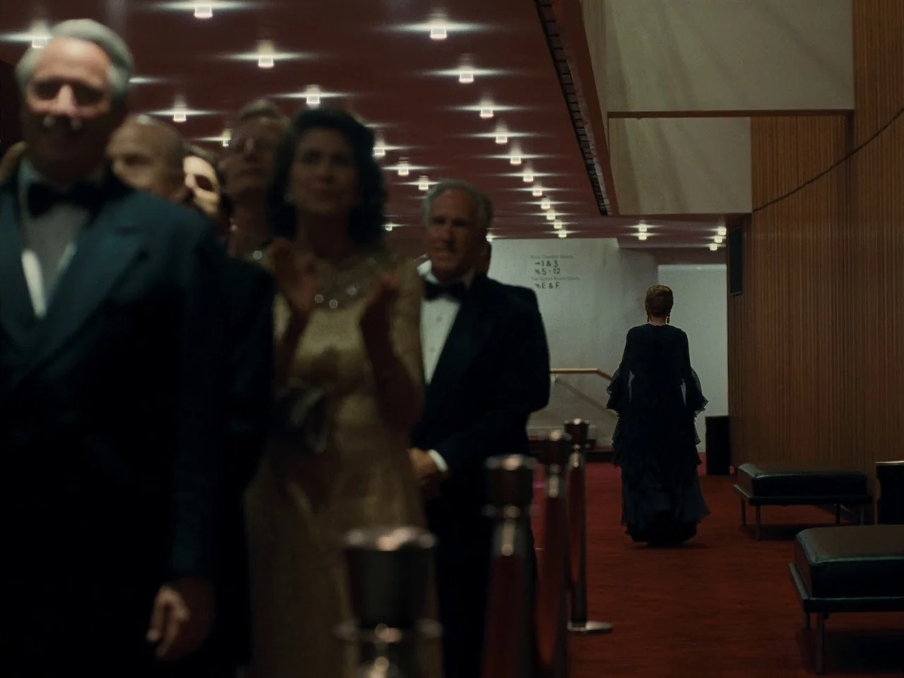 Maestro (2023) movie still: medium — A long, red-carpeted hotel or theater corridor features wood-paneled walls and low ceiling lights. A woman in a…