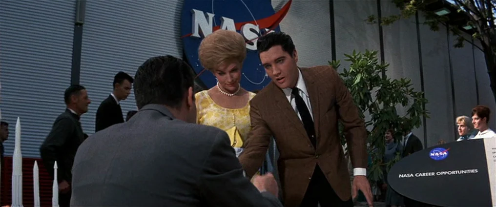 It Happened At The World’s Fair (1963) movie still: medium — An outdoor area at a NASA facility features a large wall with a logo. A man in a brown…