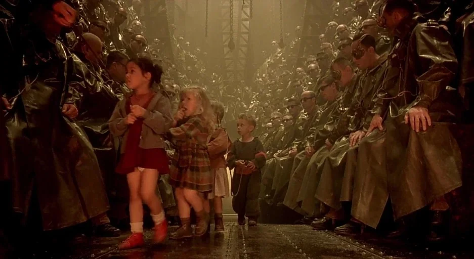 The City Of Lost Children movie still: wide — A vast, industrial interior features rows of men wearing identical dark coats and masks. Three small…