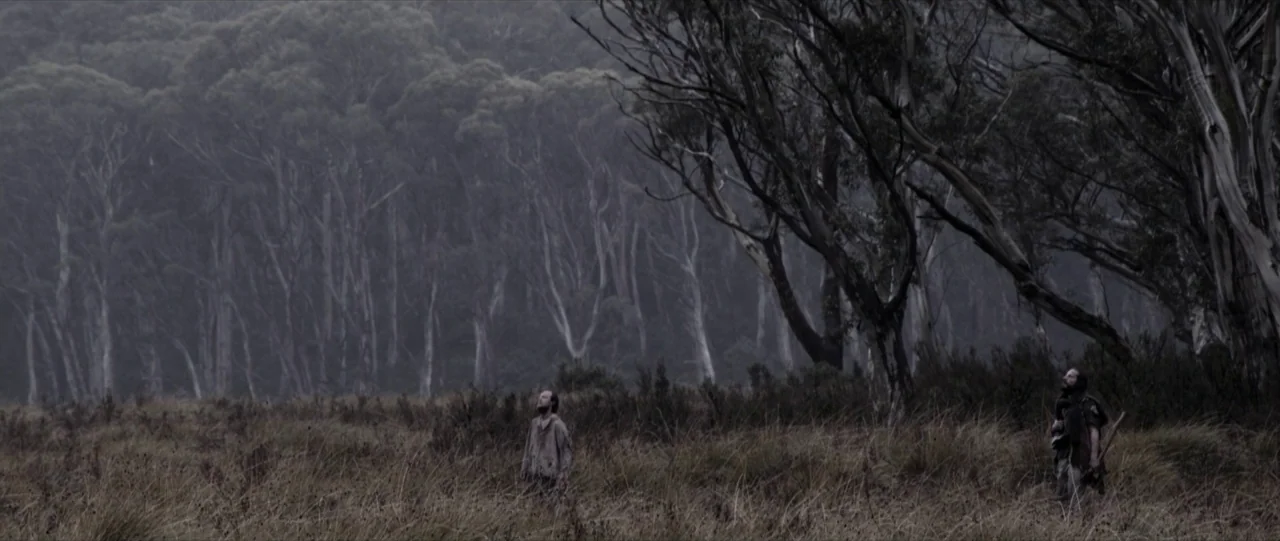 Van Diemens Land (2009) movie still: wide — A vast, open plain with tall, dry grass sits before a dense eucalyptus forest. Two men in…