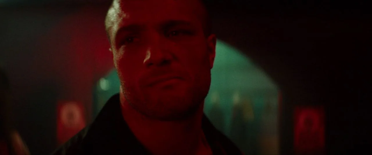 Calm With Horses (2019) movie still: closeup — A dimly lit club environment features high contrast red neon and teal background lights. A man with…
