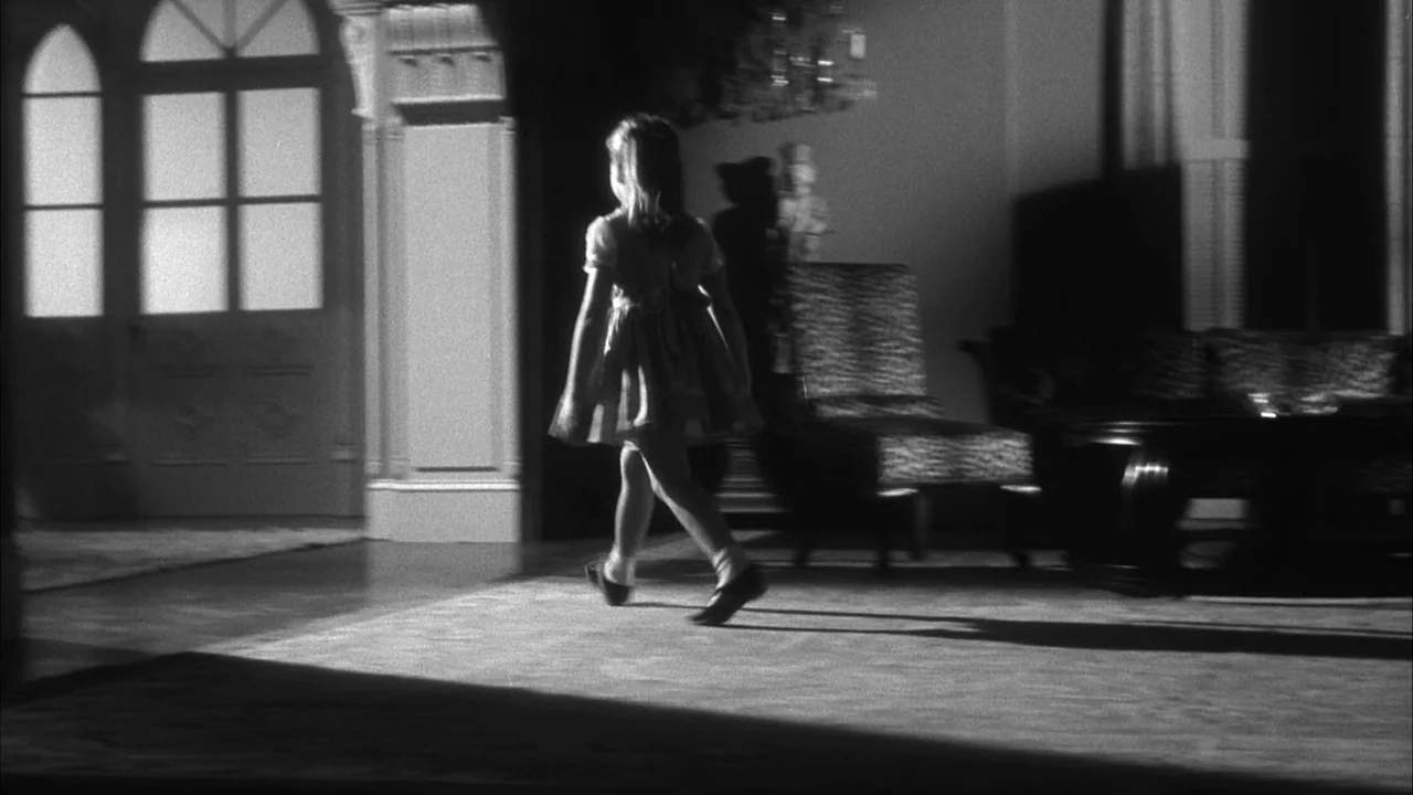 The Naked Kiss (1964) movie still: fullbody — A dark, sparsely furnished room features a large window with arched panes. A young girl moves across…