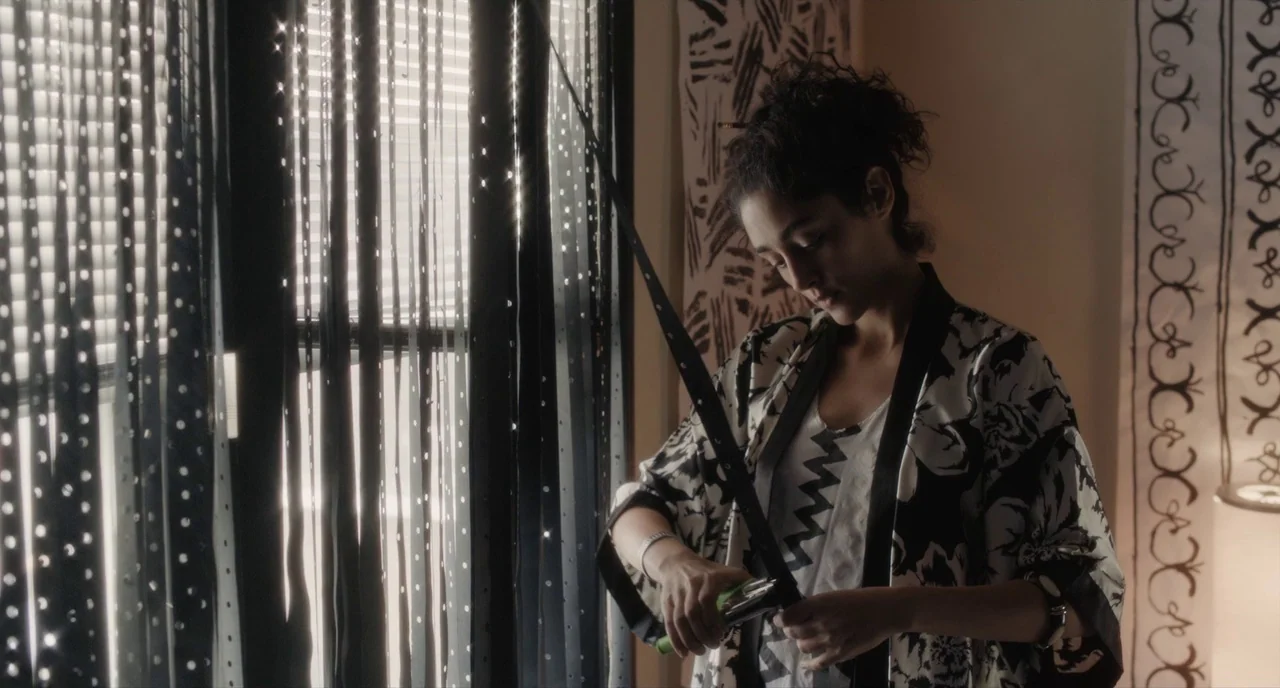 Paterson (2016) movie still: medium — A woman stands near a window with black vertical blinds casting distinct striped shadows. She holds a thin…
