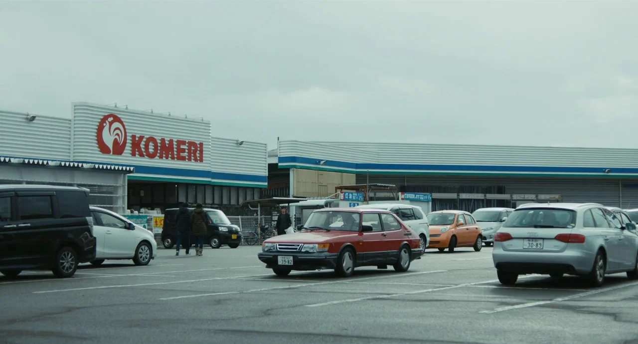 Drive My Car (2021) movie still: wide — An overcast parking lot sits in front of a white retail store with blue accents. A vintage red sedan drives…
