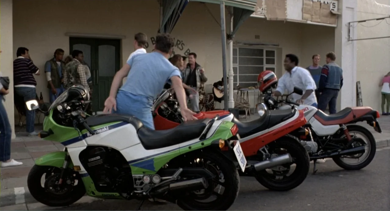 American Ninja 2: The Confrontation (1987) movie still: medium — A group of people gathers on a sidewalk in front of a shop exterior. Several 1980s…