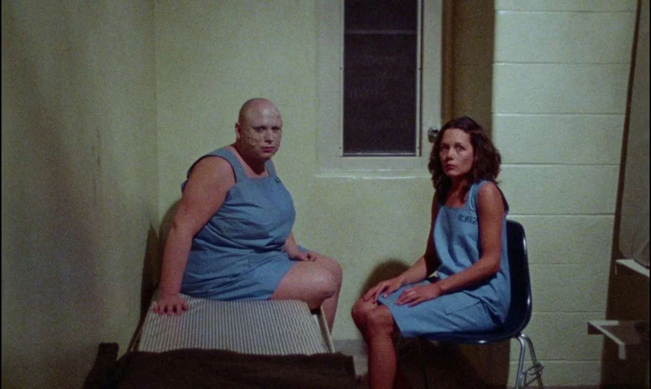 Female Trouble (1974) movie still: medium — A small room with beige concrete block walls and a barred window. A bald woman sits on a bed while a…