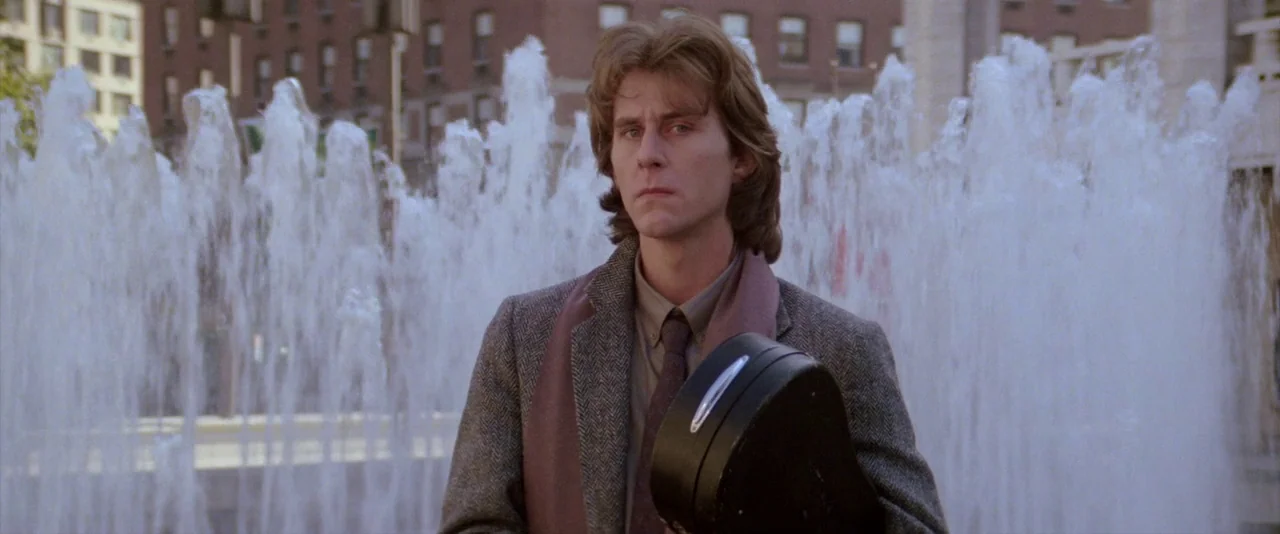 Ghostbusters (1984) movie still: medium — A man stands before a large, cascading water fountain in a paved city plaza. The man holds a hard black…