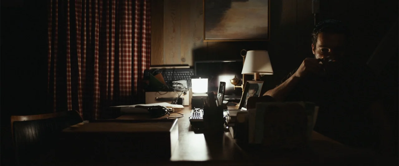 It Comes at Night (2017) movie still: medium — A dimly lit room features wood-paneled walls and a desk cluttered with electronic equipment. A man…