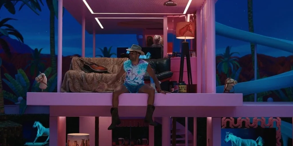 Barbie (2023) movie still: wide — A pink plastic structure resembles a life-sized dollhouse set in a dark night environment. A man sits on the floor…
