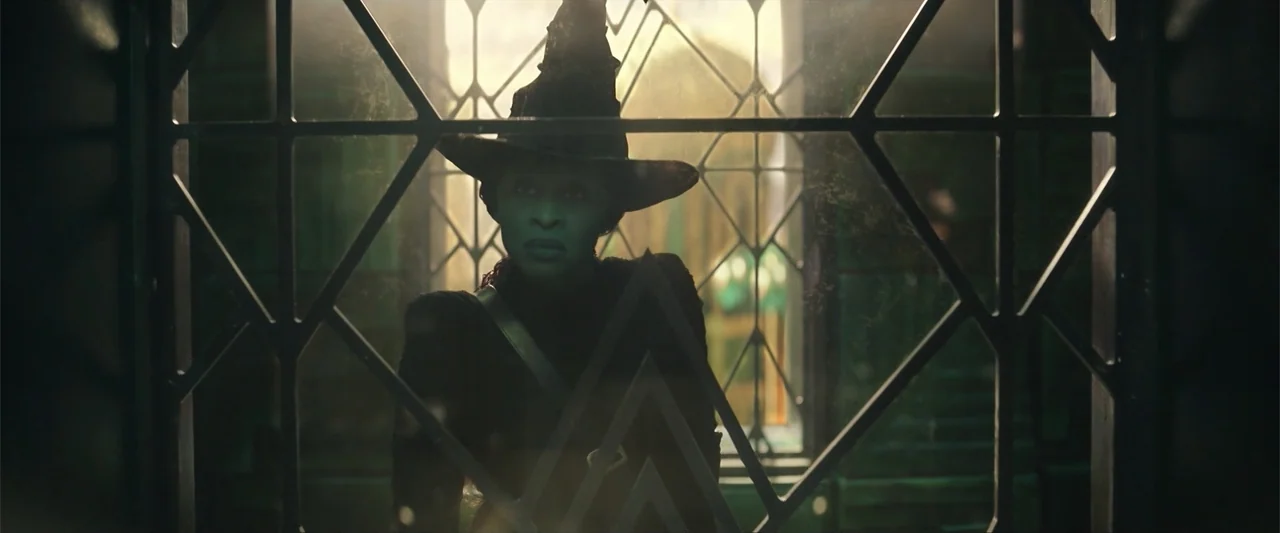 Wicked (2024) movie still: medium — An interior space viewed through a dark, decorative metal latticework window frame. A woman with green skin…