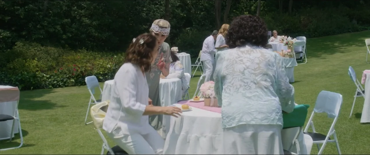 Can't Quit Mom (2022) movie still: medium — A manicured outdoor lawn is filled with small white tables set for an event. Several women wearing…