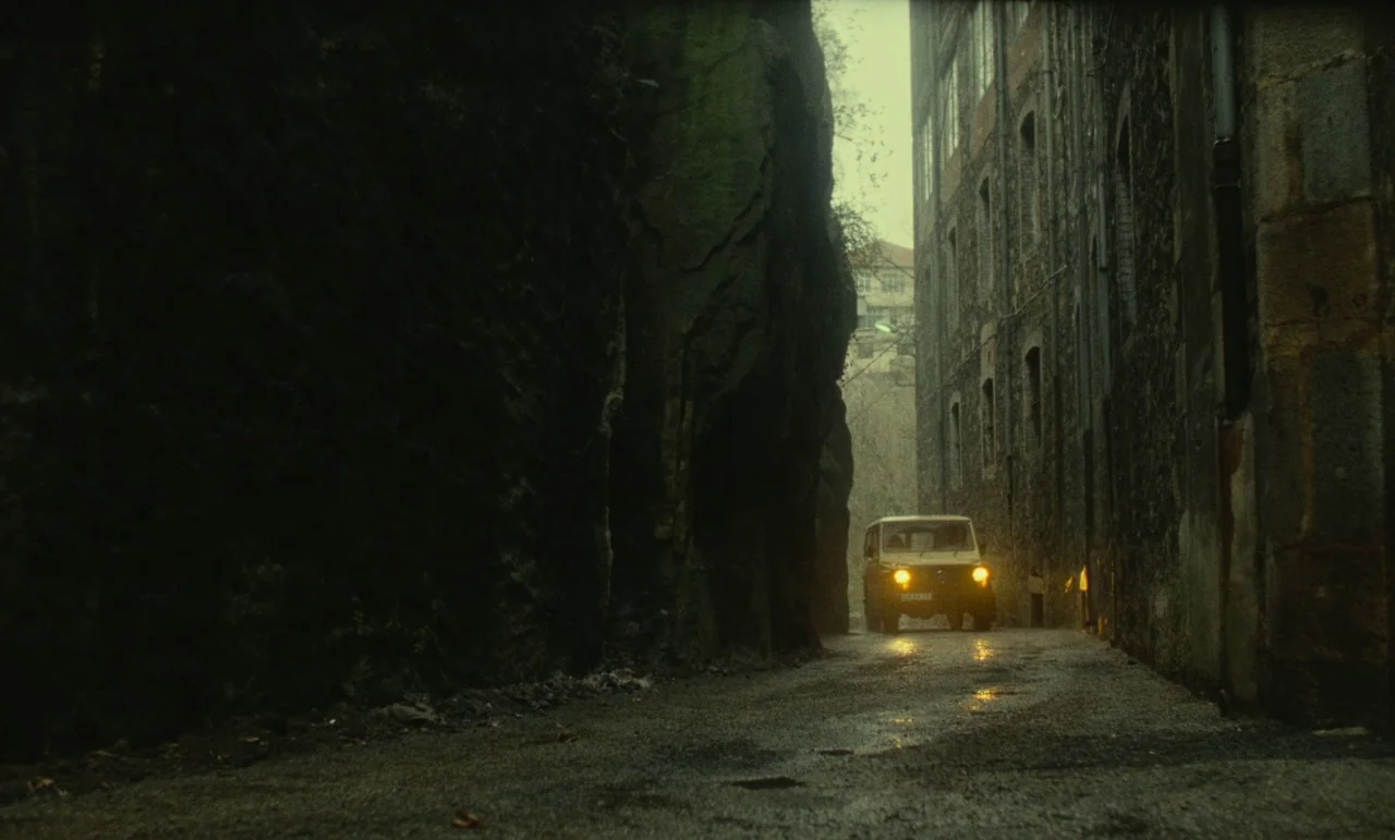 Litan (1982) movie still: wide — A narrow road is compressed between a sheer dark rock face and a weathered stone building. A beige SUV drives…