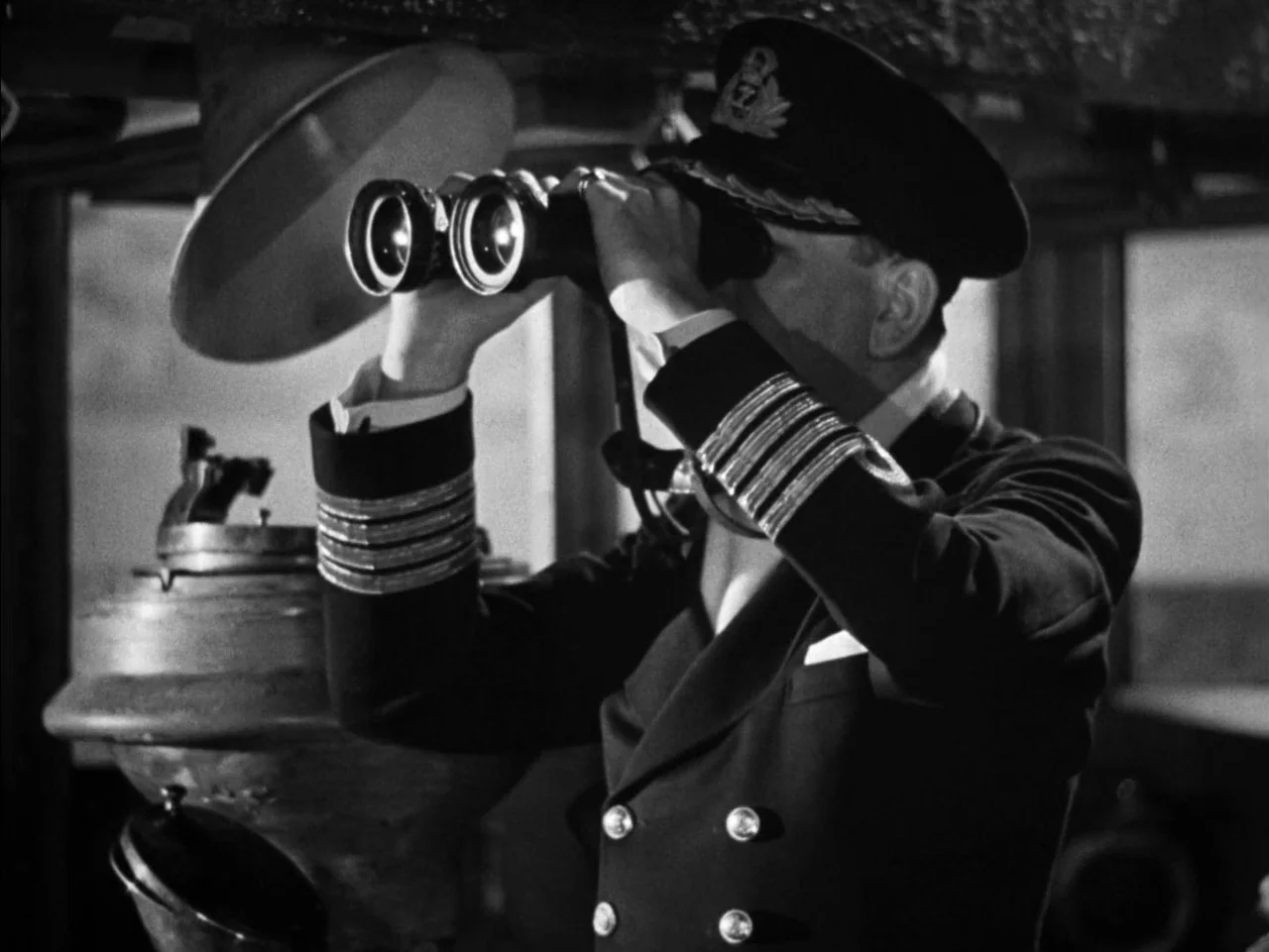 In Which We Serve (1942) movie still: medium — A dark, cramped command area on a ship with nautical equipment in the background. A naval captain in…
