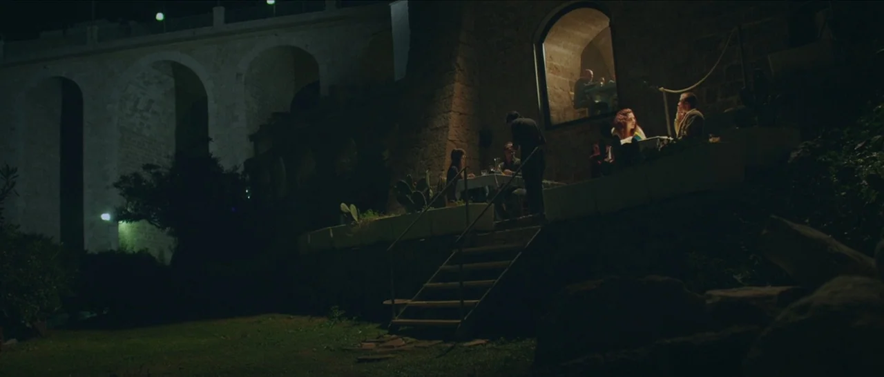 Spring (2014) movie still: wide — An outdoor restaurant terrace sits at the base of a large stone arched bridge. Several people eat at tables under…