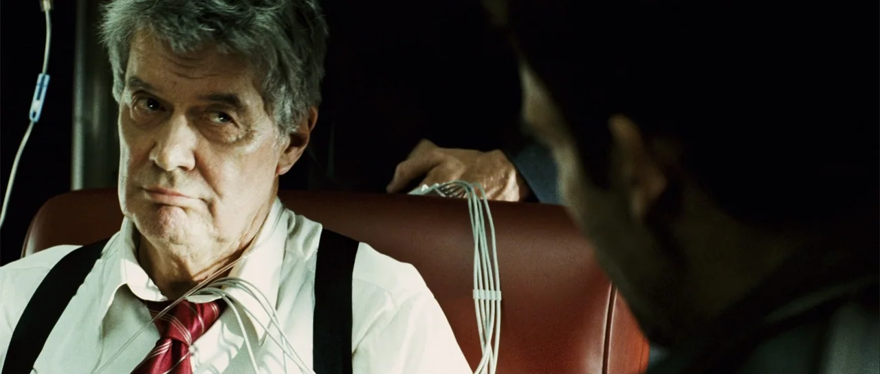 Shoot 'Em Up (2007) movie still: closeup — A dark, spartan room features a red leather chair and diagnostic testing equipment. An older man wearing…
