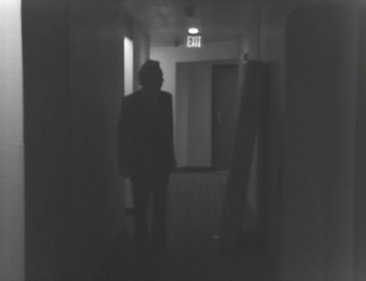 Computer Chess (2013) movie still: fullbody — A dimly lit hotel corridor features a glowing overhead exit sign in the distance. A man in a dark suit…