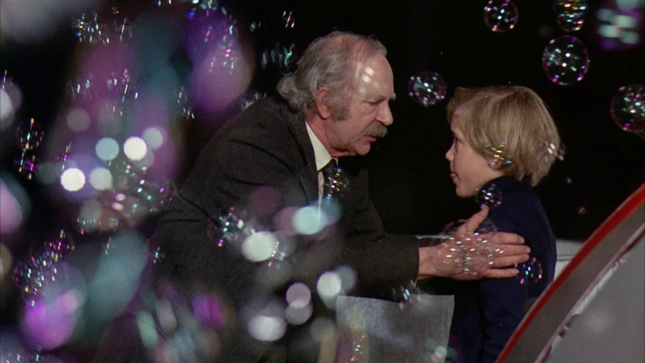 Willy Wonka & the Chocolate Factory (1971) movie still: medium — An older man in a suit faces a young boy against a dark backdrop. Multiple bubbles…
