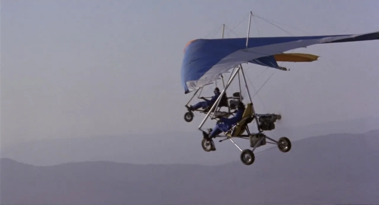 American Ninja 3: Blood Hunt (1989) movie still: wide — Two motorized hang gliders drift through the clear blue sky over distant mountains. Each…