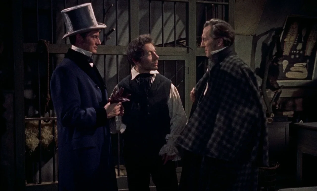 The Revenge of Frankenstein movie still: medium — A dimly lit room features a metal grate backdrop and wooden furniture. Three men in period…