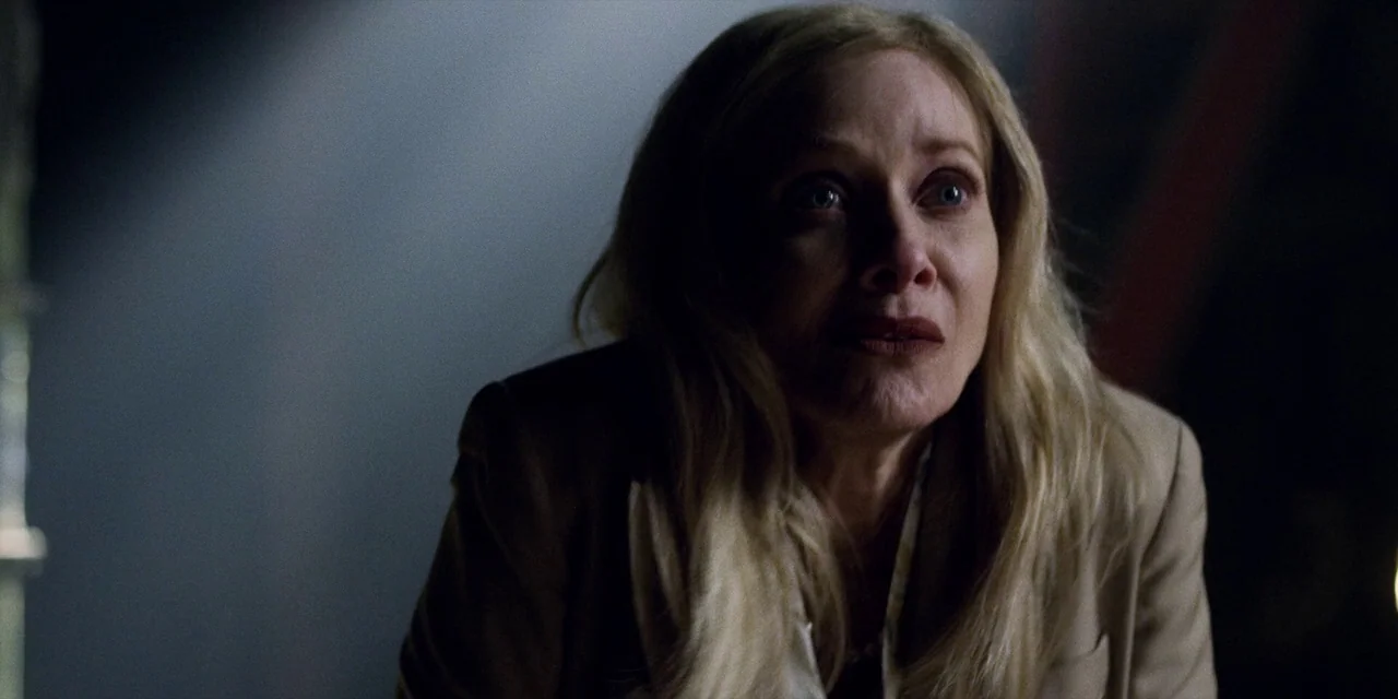Jakob’s Wife (2021) movie still: closeup — A dimly lit interior space features a blonde woman in a tan blazer. She looks upward with visible strain…