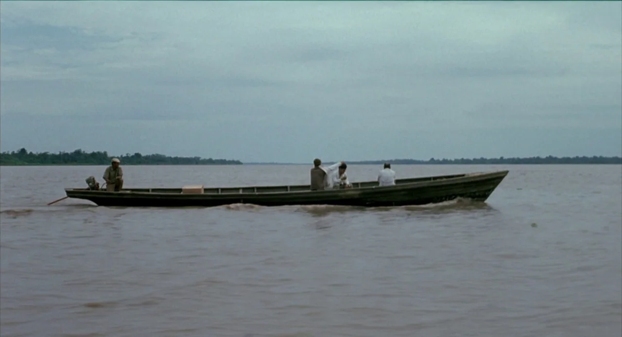The Motorcycle Diaries (2004) movie still: wide — A long, narrow wooden boat moves across a wide, murky river under an overcast sky. Four men sit…