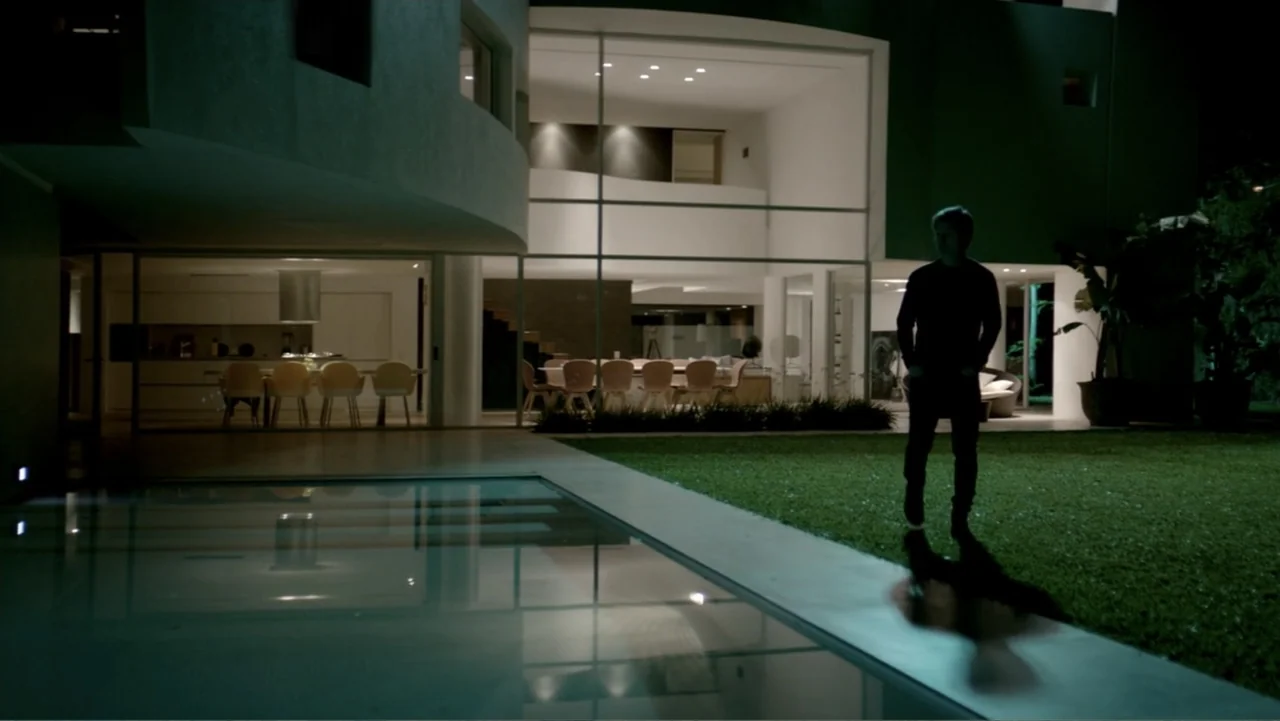 I Married a Dumbass (2016) movie still: fullbody — A modern residence with large glass walls stands adjacent to a dark swimming pool. A man walks…