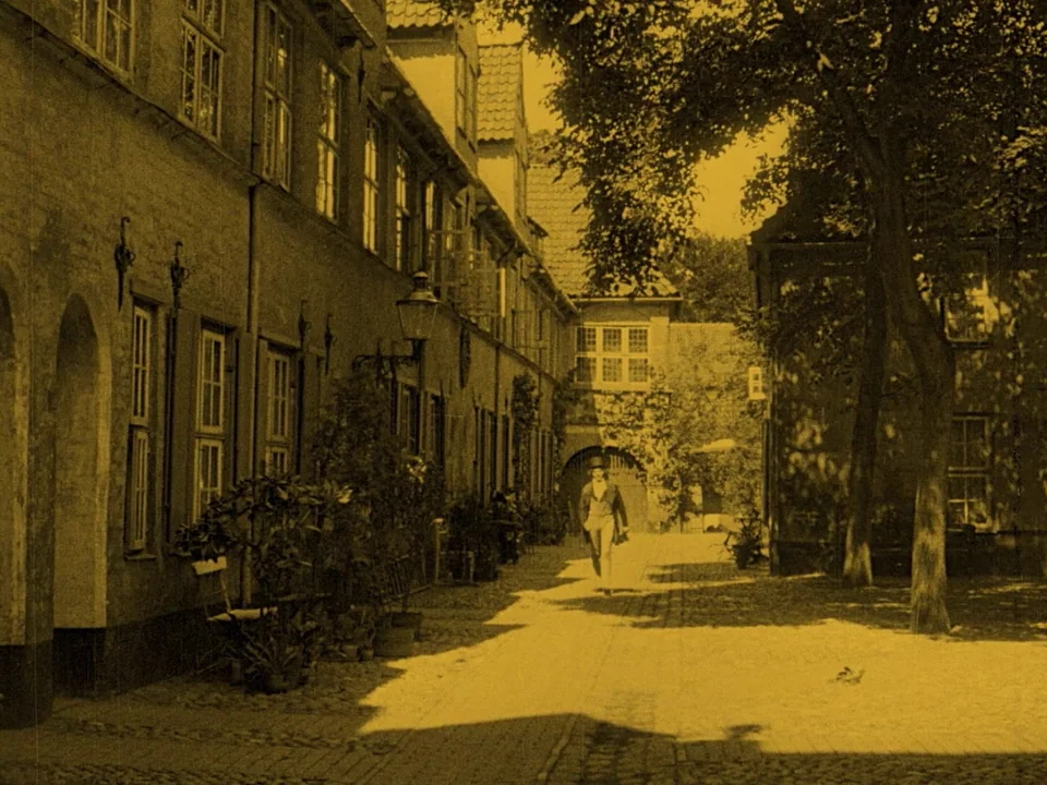 Nosferatu (1922) movie still: wide — A sunlit stone courtyard is flanked by old multi-story brick buildings with large windows. A man wearing a top…