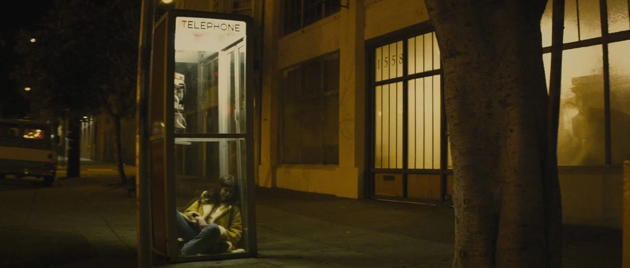 The Diary of a Teenage Girl (2015) movie still: wide — A nighttime sidewalk view features a lit telephone booth adjacent to a large tree. A woman…