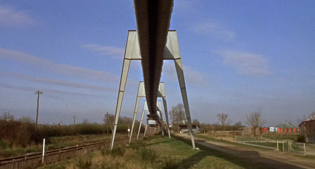 Fahrenheit 451 (1966) movie still: establishing — A metal monorail structure spans above a rural area with grass and dirt paths. A small train…