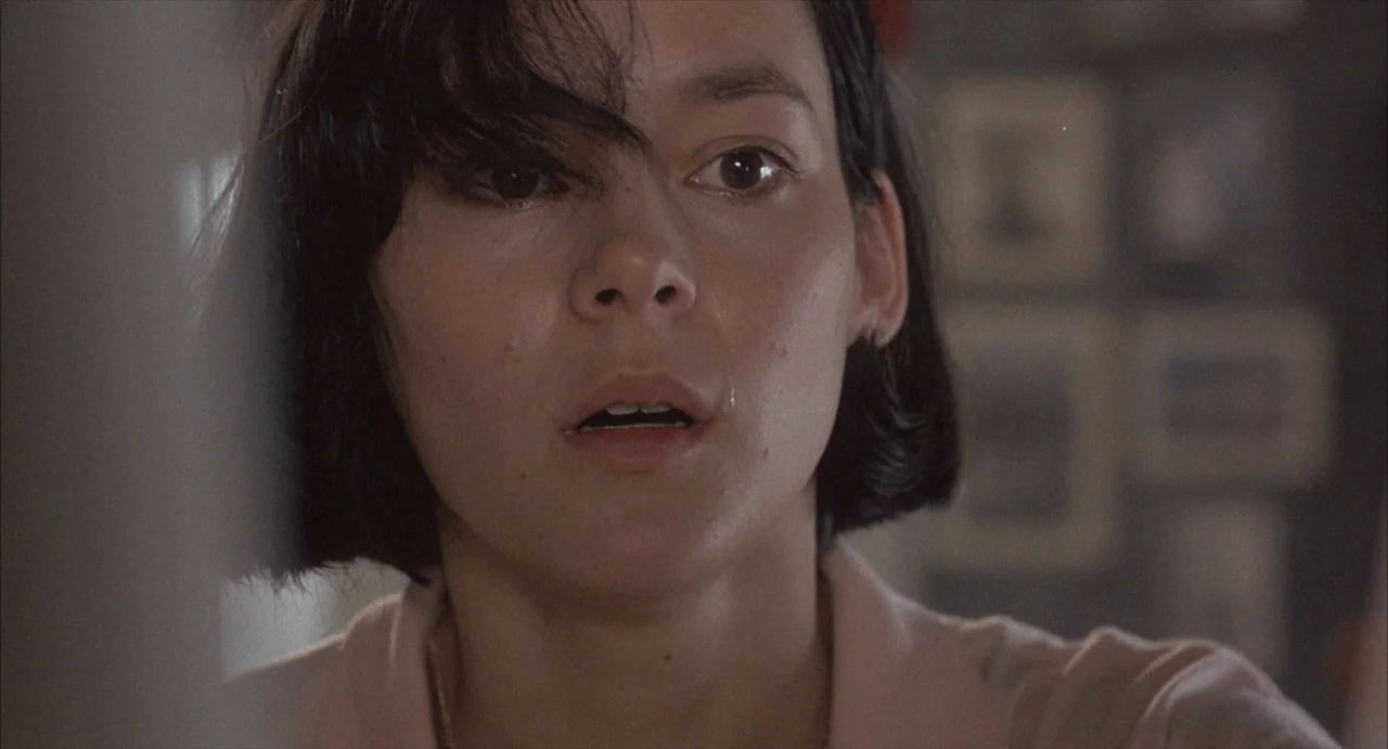 Masquerade (2023) movie still: closeup — An interior space contains a person in front of a blurred wall with frames. A woman with short black hair…