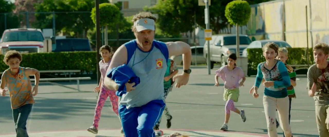 Cooties (2014) movie still: fullbody — An asphalt playground serves as a running space for a man and multiple children. Several children have pale,…
