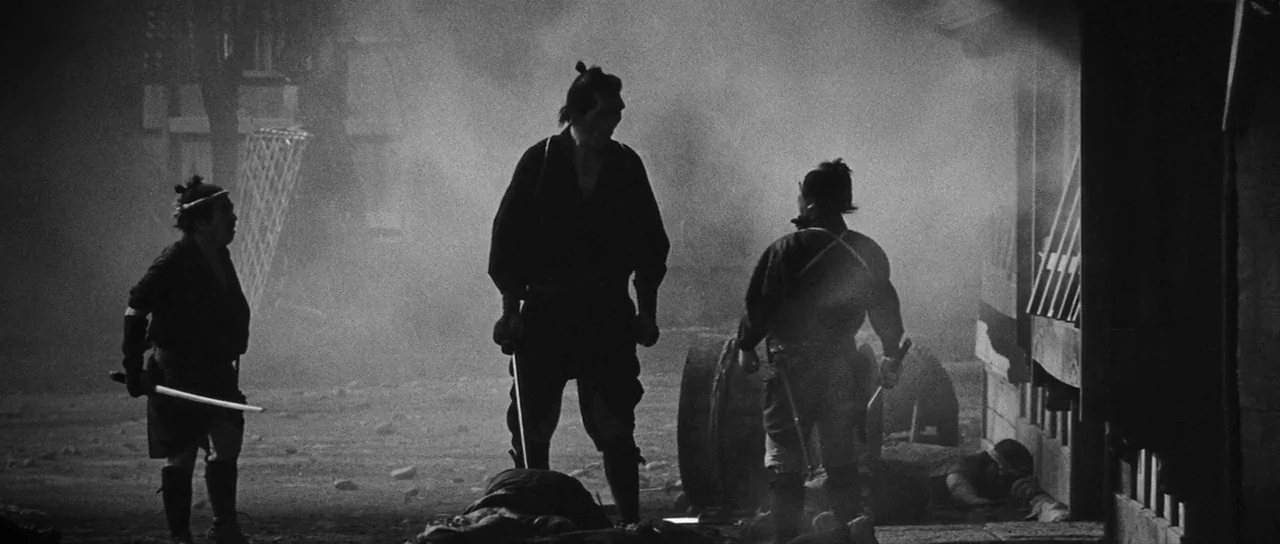Yojimbo (1961) movie still: fullbody — A dark, foggy outdoor village area at night with wooden architecture along the side. Three men standing with…