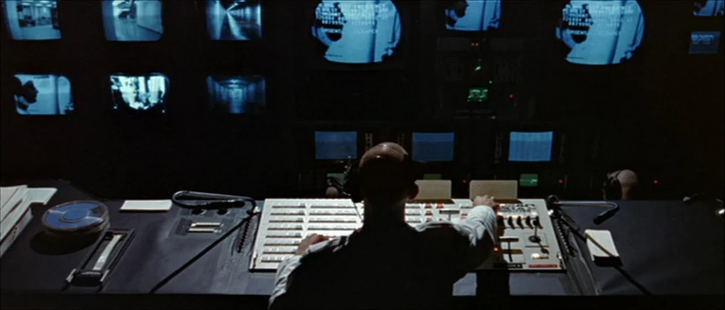 THX 1138 (1971) movie still: medium — A dimly lit room is filled with a wall of active security camera monitors. A man sits at a long desk operating…