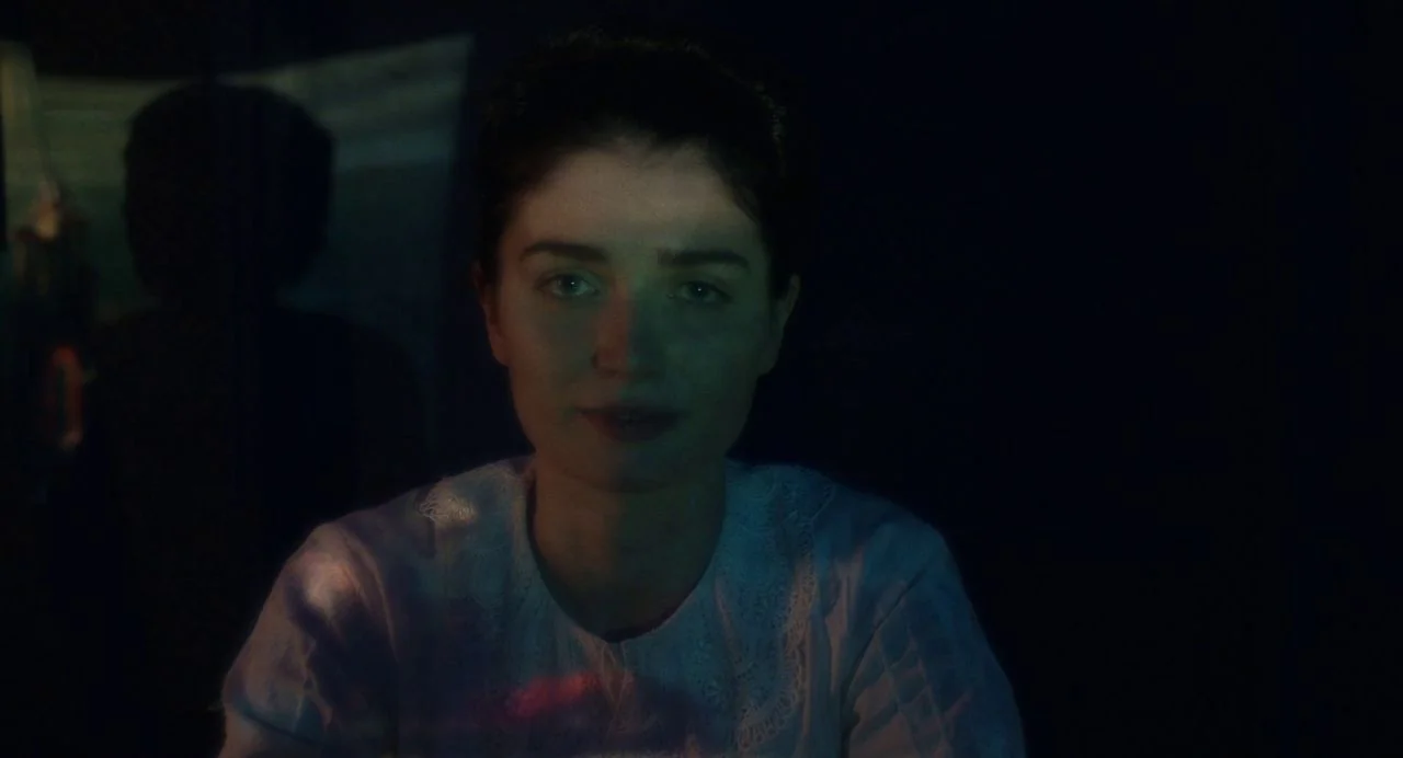 Tesla (2020) movie still: closeup — A person sits in a darkened room with no background details visible. They look directly at the lens while…