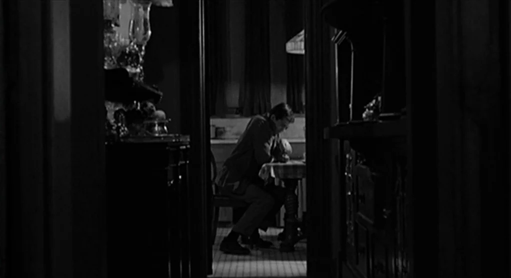 Psycho movie still: wide — The frame views a dim room through a narrow, dark door frame. A man sits at a small table while resting his head on his…