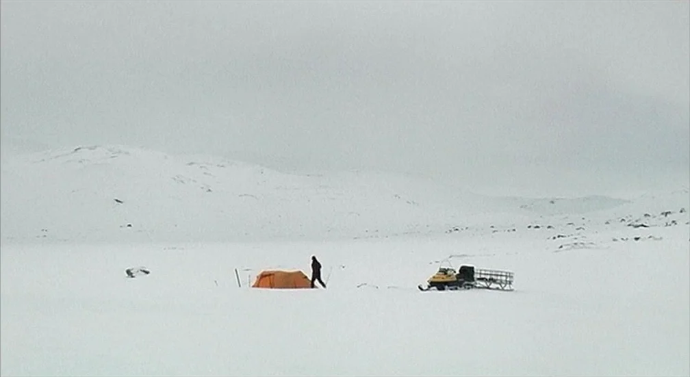 9 Songs (2004) movie still: wide — A vast, flat, snow-covered tundra expanse under a uniform grey overcast sky. A person walks from a small orange…